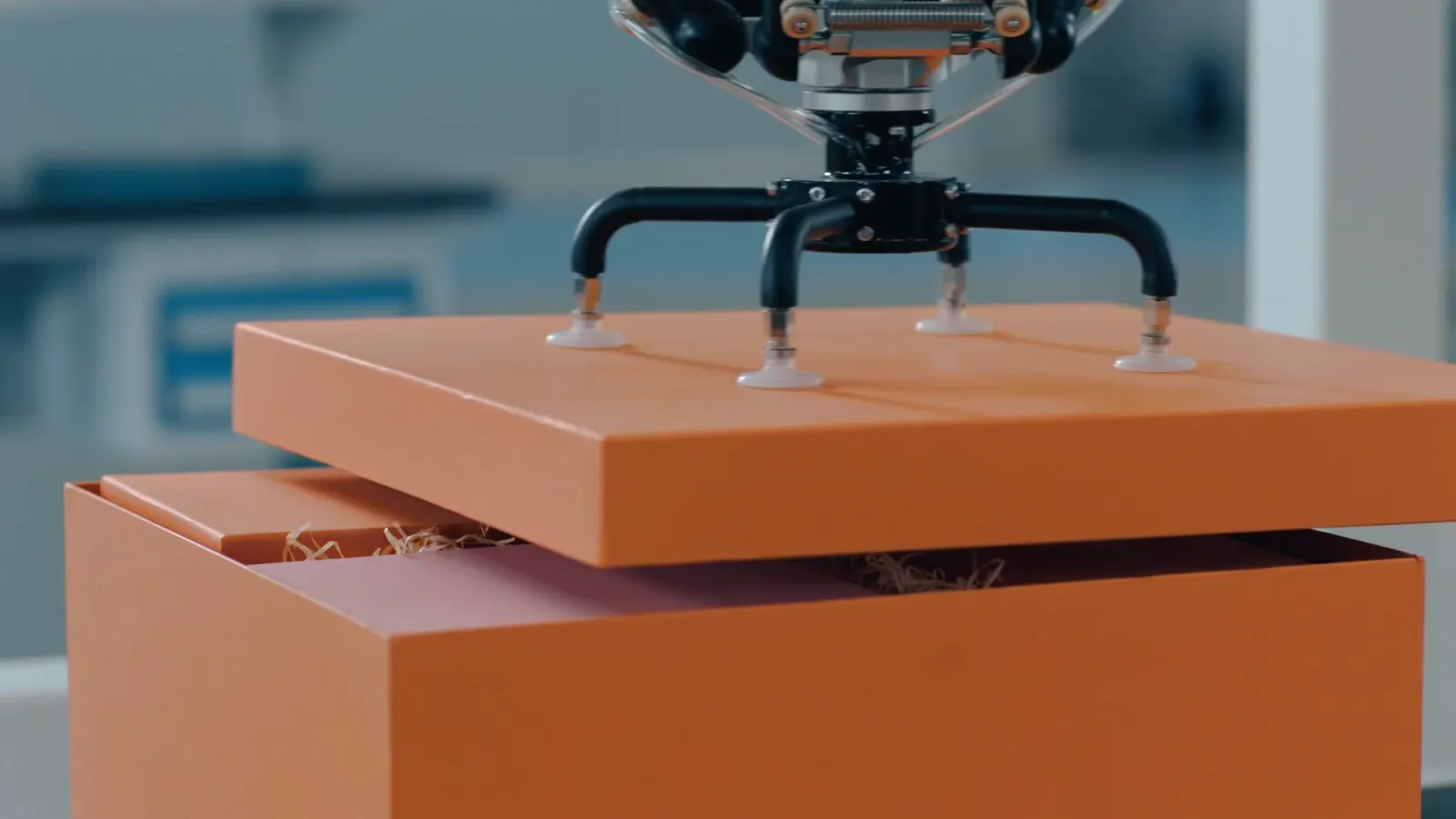 白い多関節ロボットのアームと、オレンジ色の箱が並ぶ作業台
